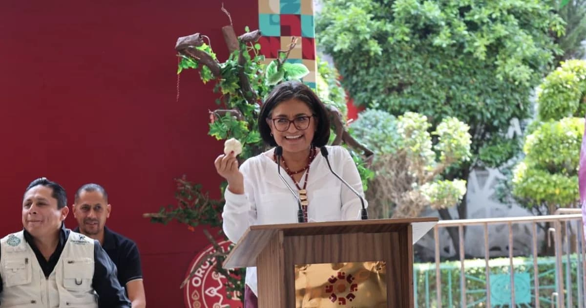 Una mujer habla desde un podio durante un evento de la Alcaldía Iztapalapa, con dos hombres sentados a un costado y vegetación al fondo.