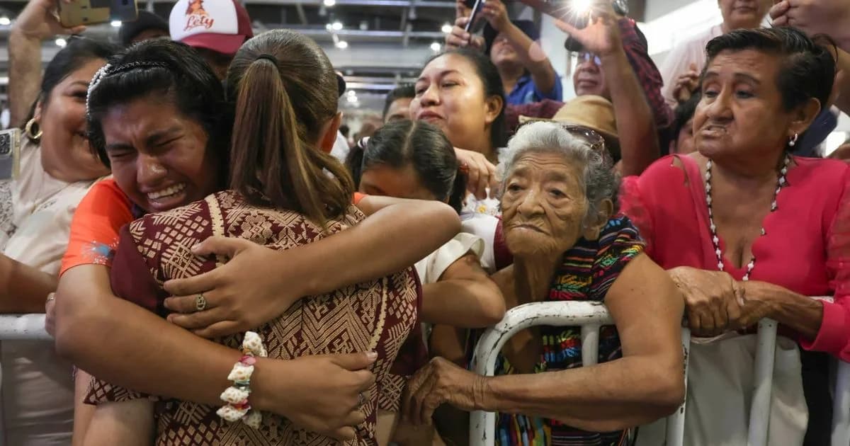 Claudia Sheinbaum abraza a una mujer entre asistentes a un acto público, mientras varias personas observan y toman fotografías.