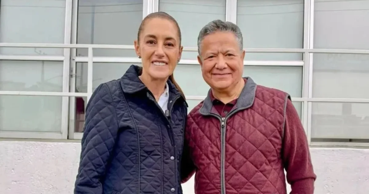 Dos personas sonrientes, una mujer con chamarra azul marino y un hombre con chaleco guinda, frente a una fachada de vidrio