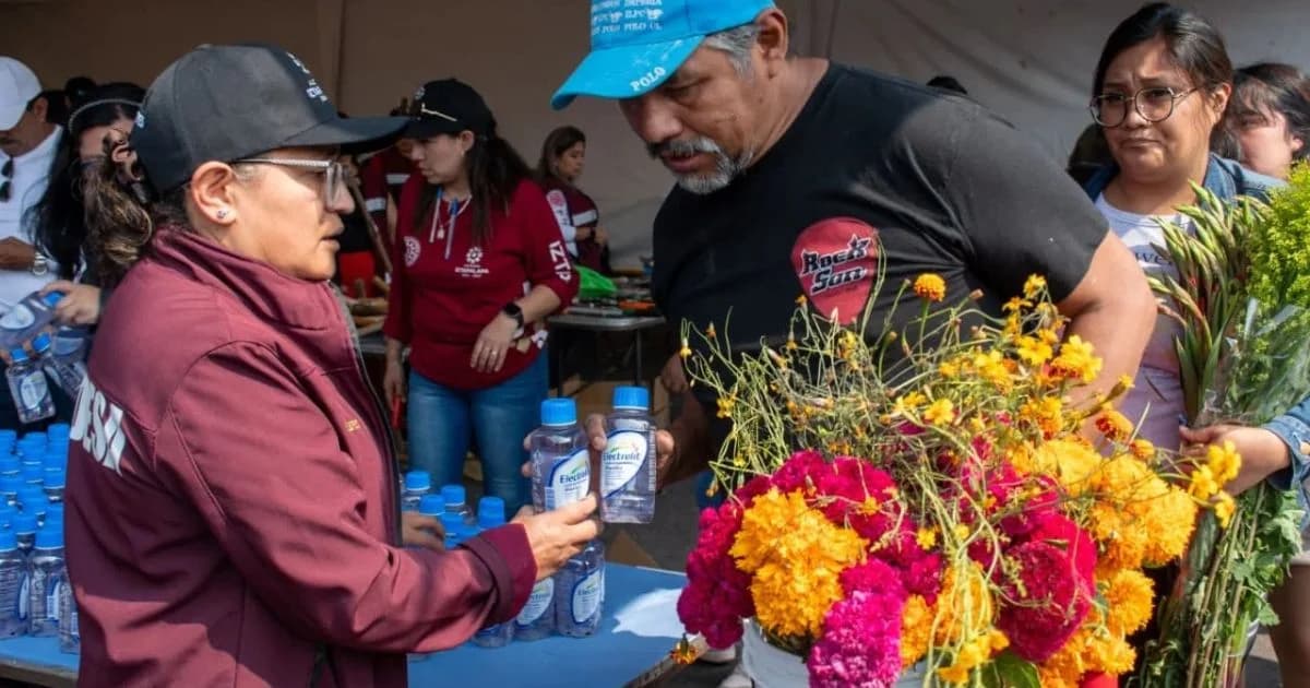 Una mujer entrega botellas de agua a un hombre que sostiene flores de cempasúchil, junto a una mesa de hidratación.