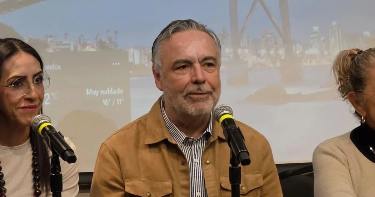 Participantes en una mesa de conferencia con micrófonos; al centro, un hombre de chaqueta café