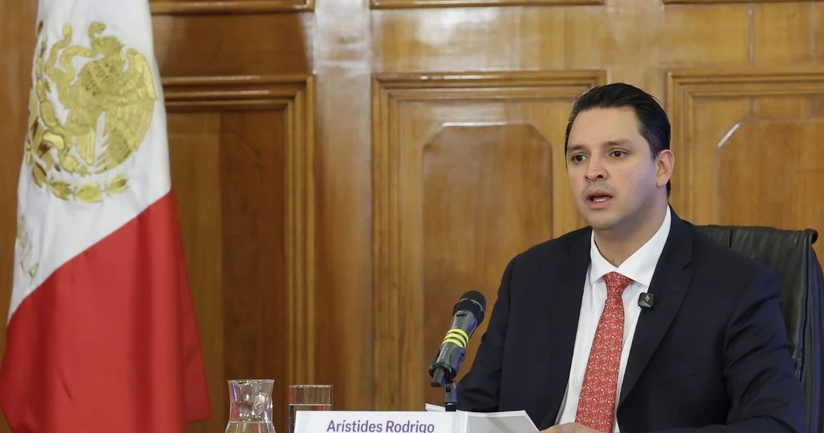 Arístides Rodrigo Guerrero García interviene ante un micrófono junto a la bandera de México