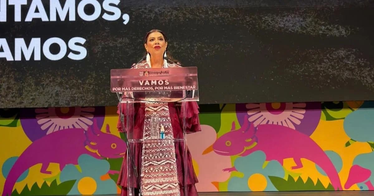 Mujer habla desde un atril en un escenario con la frase Vamos por más derechos, por más bienestar