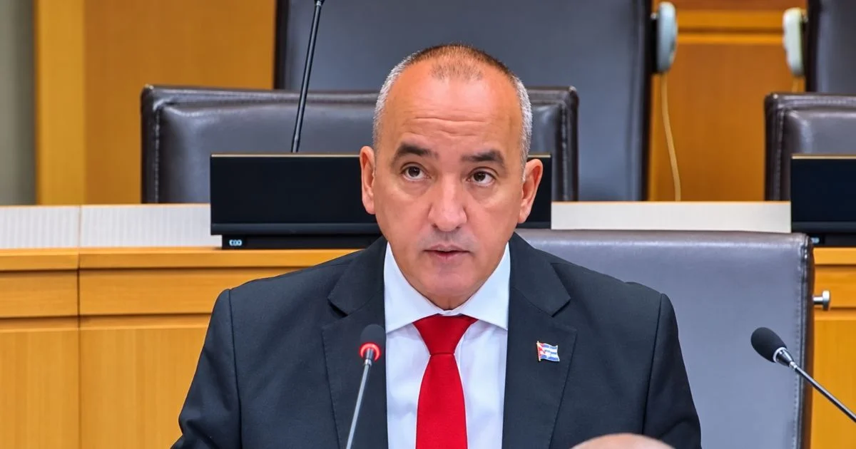 Diplomático de traje oscuro y corbata roja interviene ante micrófonos en una sala de reuniones.