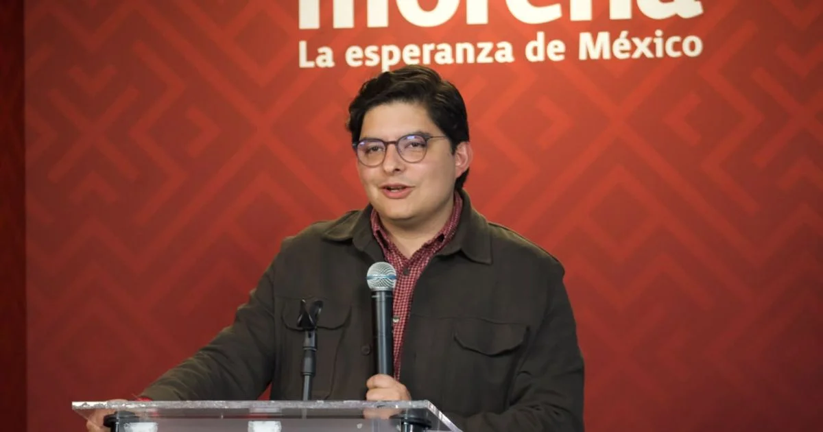 Hombre con lentes habla ante un micrófono y un atril, con el logotipo de Morena al fondo