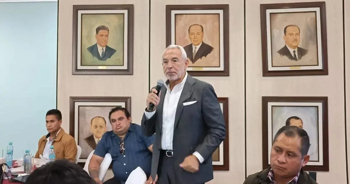Hombre con traje sostiene un micrófono durante una reunión en un salón con varias personas sentadas