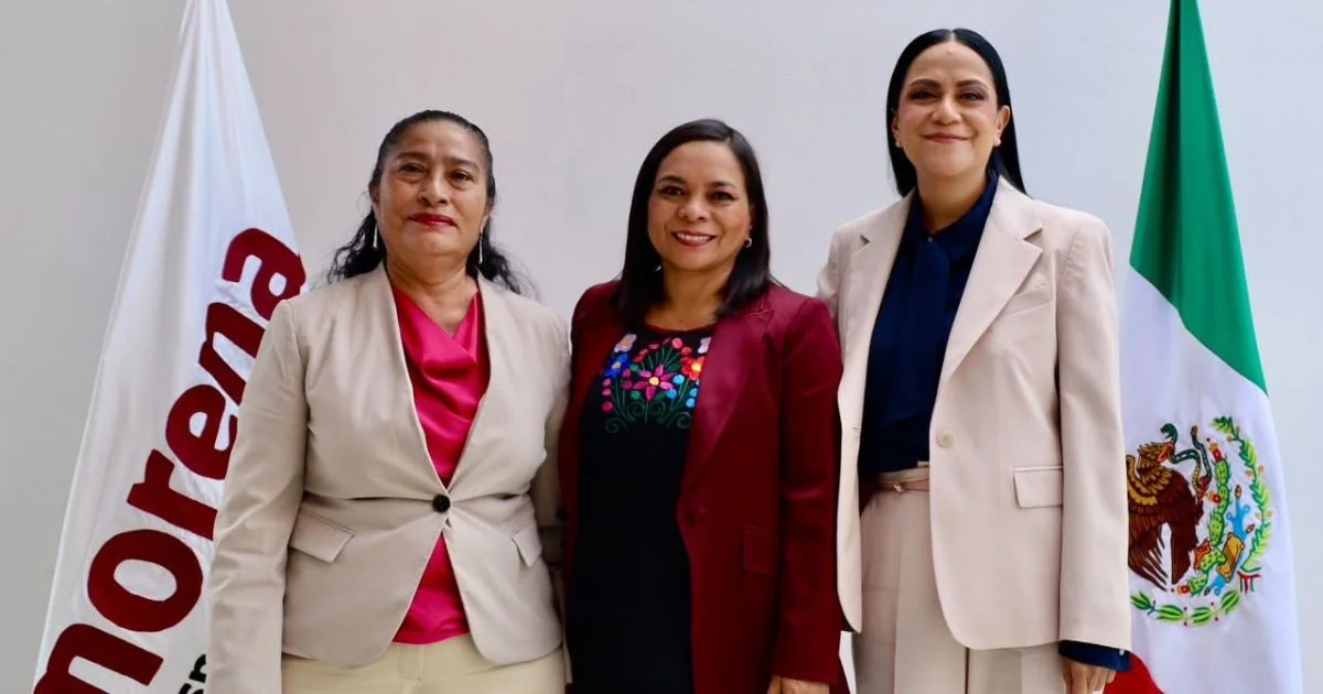 Tres mujeres posan juntas frente al emblema de Morena, entre una bandera del partido y la bandera de México.