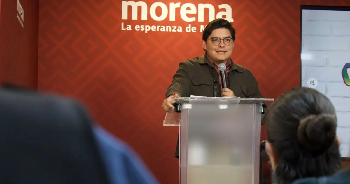 Un hombre con lentes habla ante un micrófono detrás de un atril transparente, frente a un fondo rojo con el logotipo de Morena y asistentes en primer plano